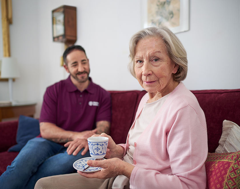 Leben mit Demenz - Mitarbeiter von Home Instead mit einer älteren Kundin, die im Vordergrund des Bildes ist und eine Tasse Tee in der Hand hält